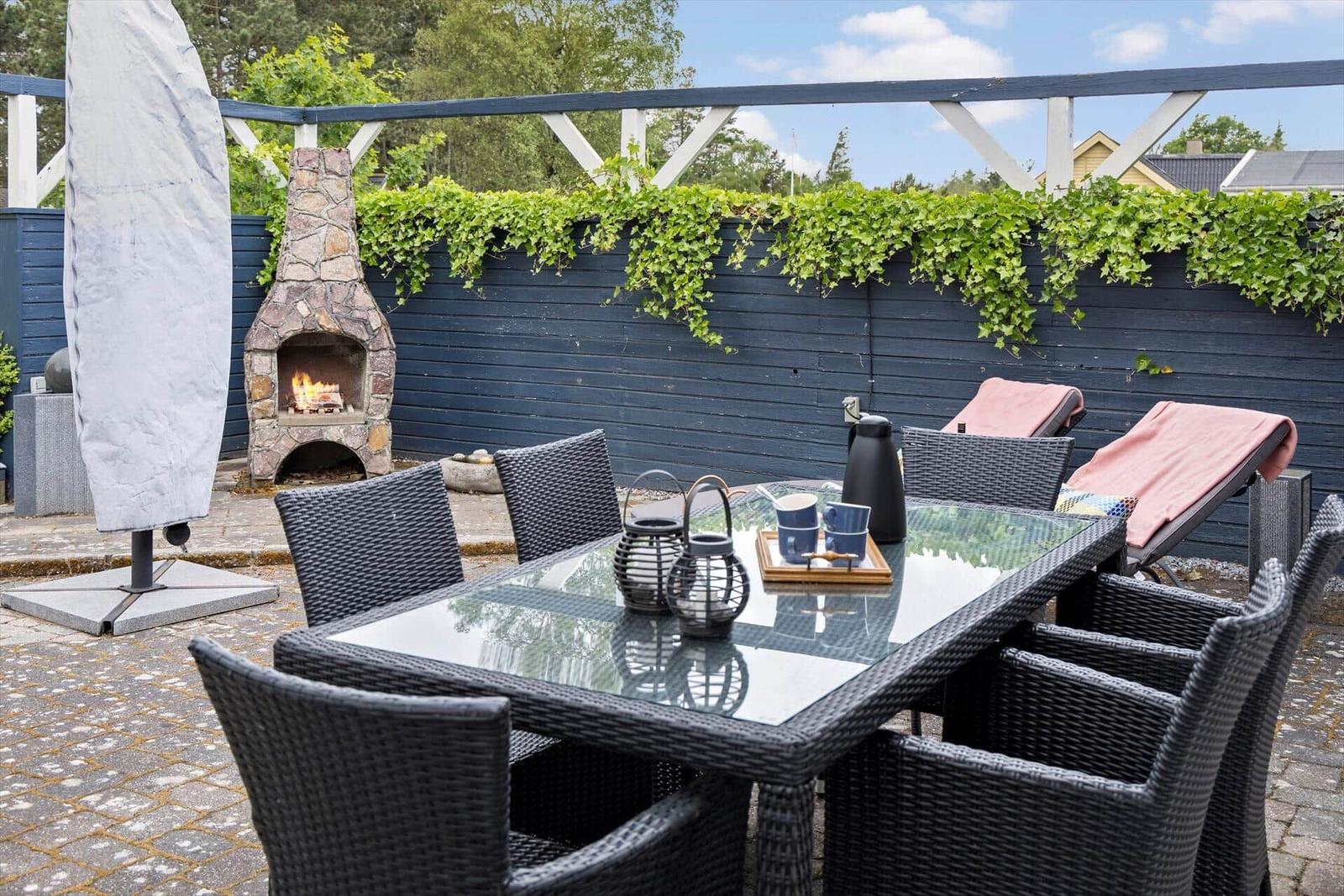 Terrace with table, chairs, and stone fireplace. Background: dark blue fence with ivy.