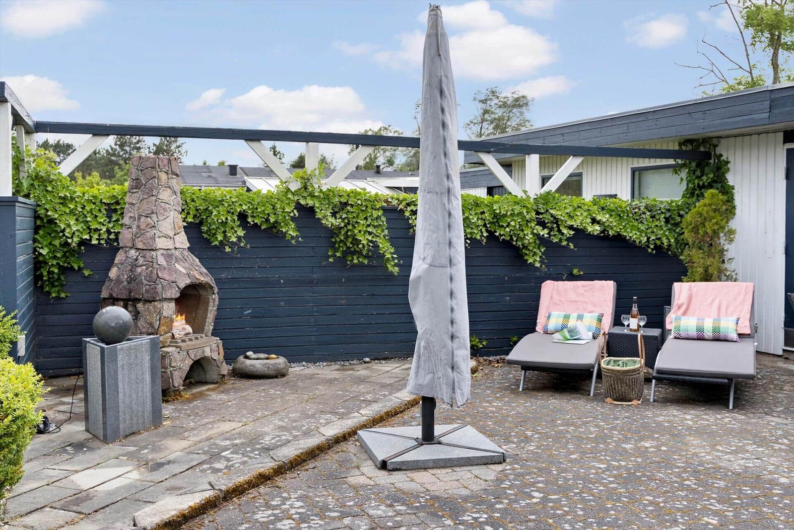 Terrace with loungers, umbrella, and stone fireplace. Background: dark fence with ivy.