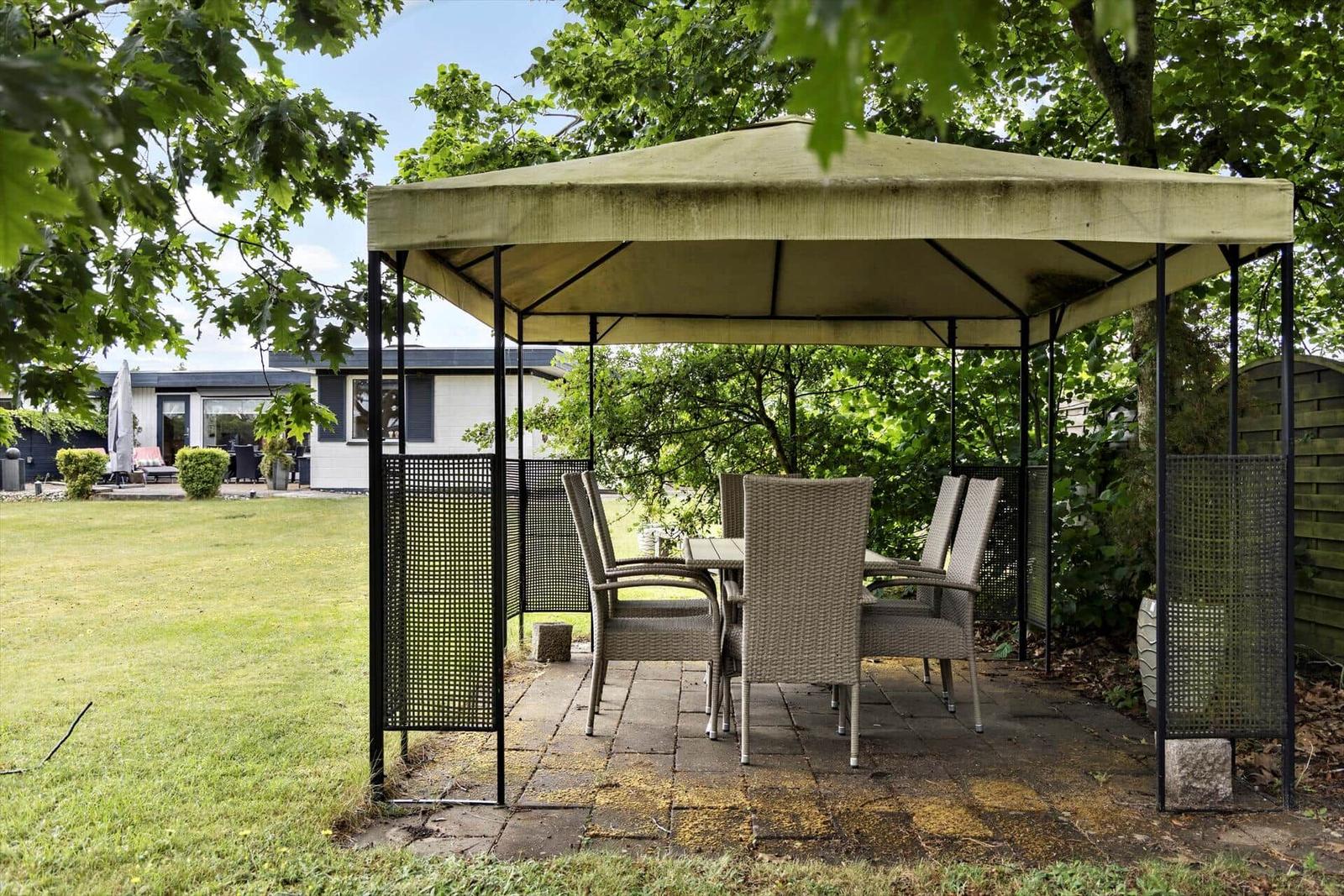 Garden with pavilion, table and chairs on stone slabs.