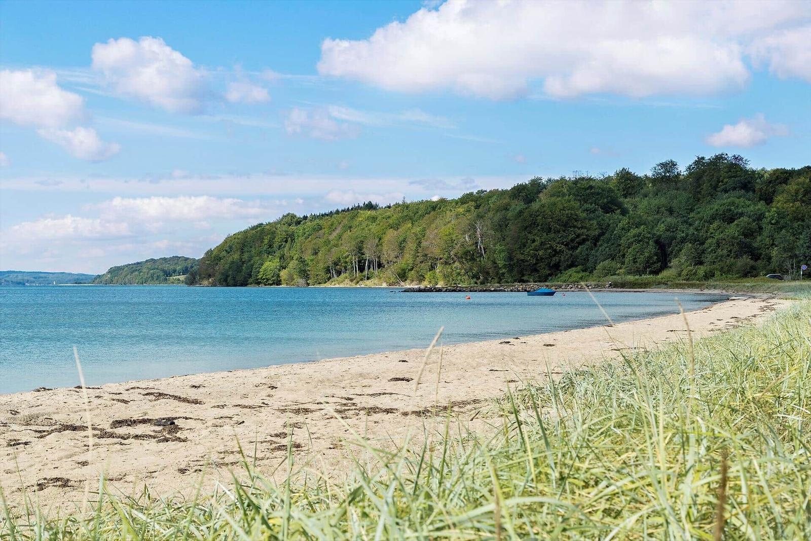 Strand mit Sand, Gras und blauem Wasser. Wald im Hintergrund. Ein kleines Boot liegt im Wasser.