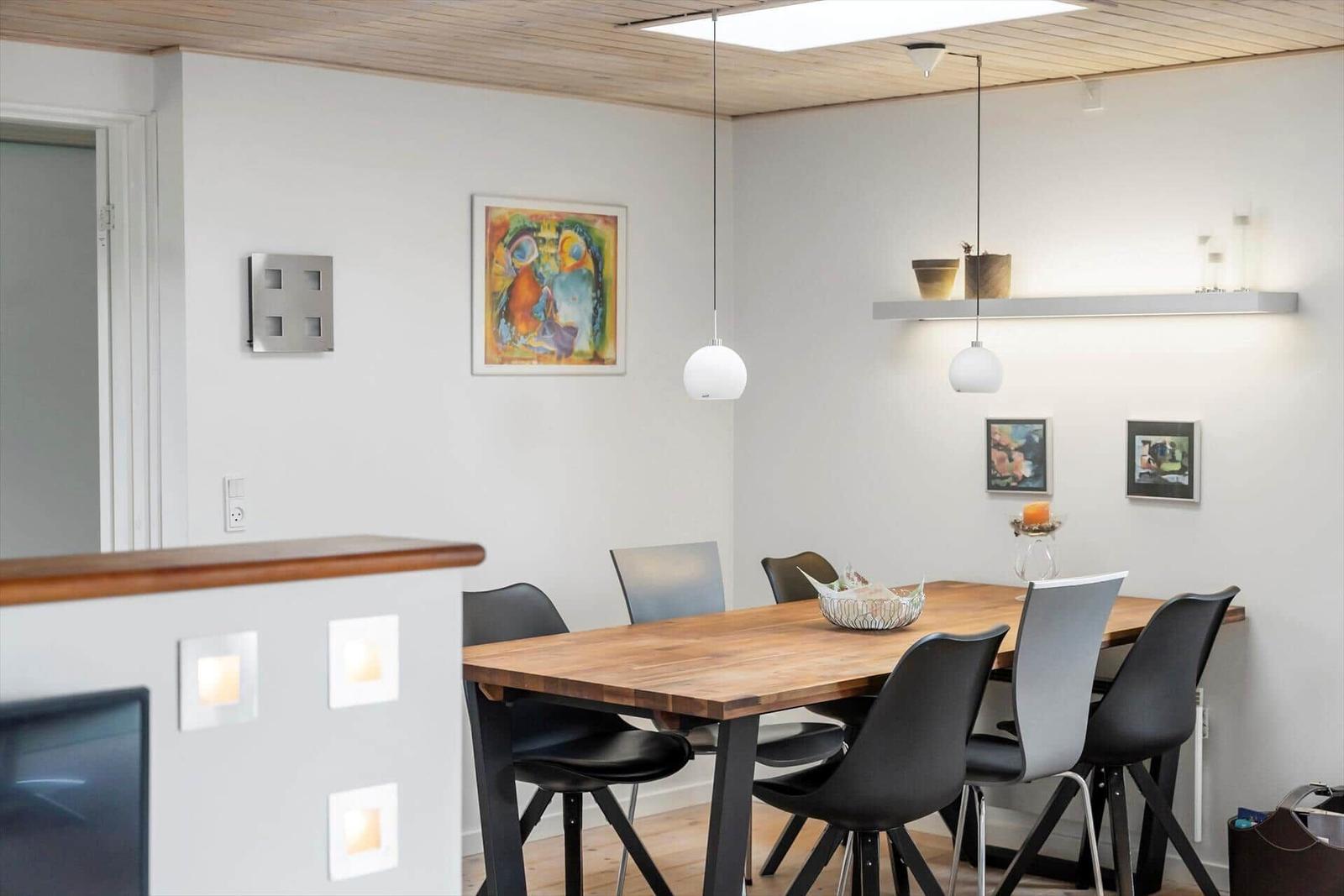 Esszimmer mit Holztisch, Stühlen und Wandbildern. Deckenleuchten und Regal mit Pflanzen.