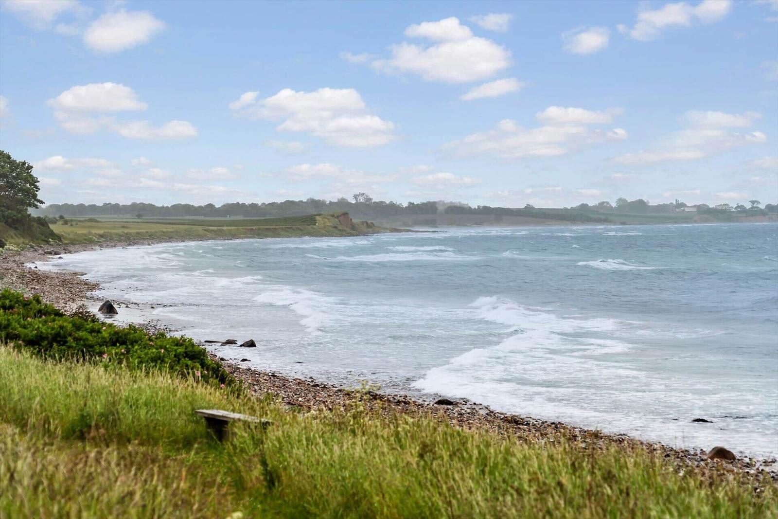 Strand mit Steinen und Gras, Wellen brechen sanft an Küste.