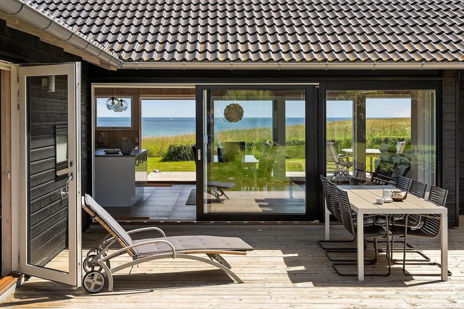 Terrasse mit Liegestuhl und Tisch. Durchgehende Glasfront mit Blick auf Strand und See.