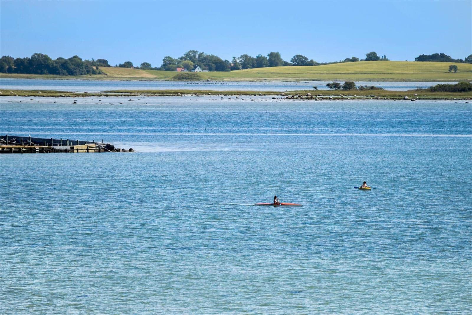 Zwei Personen paddeln auf ruhigem Wasser. Im Hintergrund sind Felder und Bäume zu sehen.