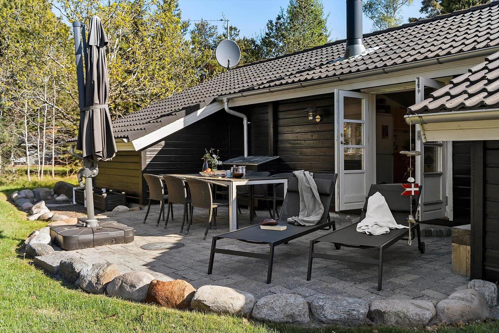 Terrasse mit Tisch, Stühlen und Liegen vor einem Holzhaus mit offener Tür.