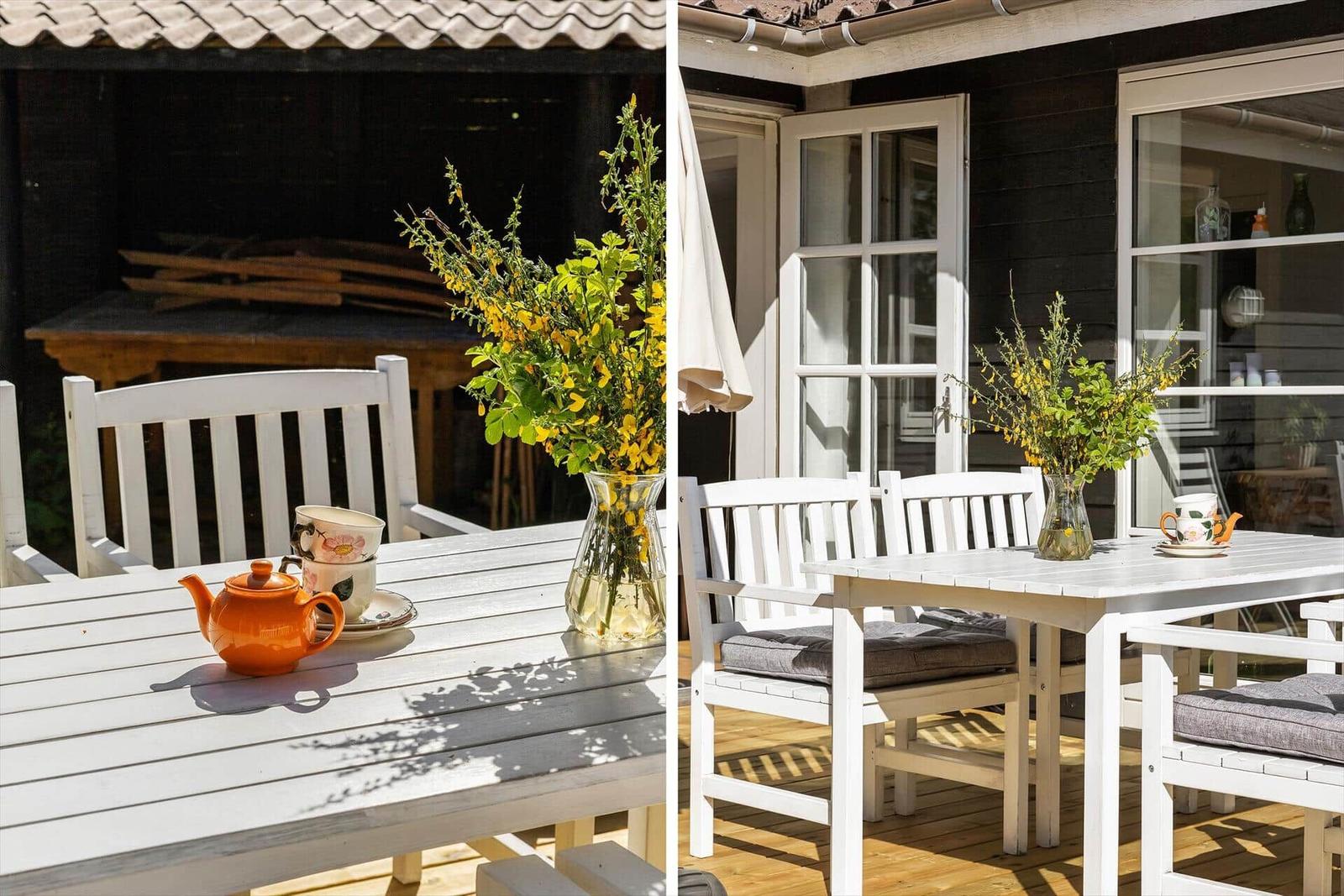 Weiße Terrassenmöbel mit Teekanne und Blumen auf Holzterrasse vor Haus.