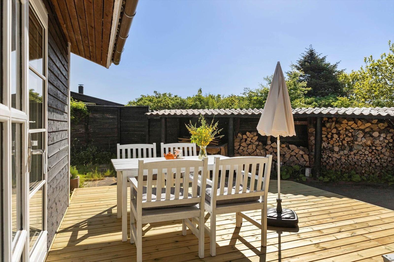Terrasse mit weißem Tisch und Stühlen, Sonnenschirm und Holzstapel im Hintergrund.