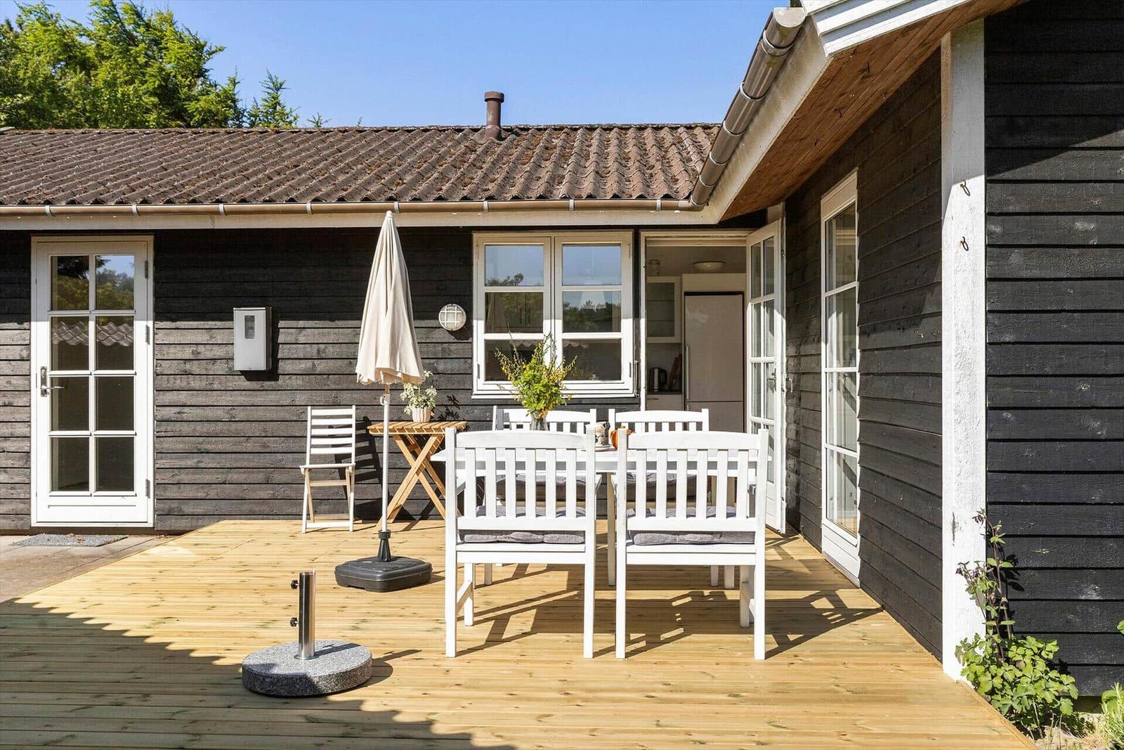 Terrasse mit weißem Tisch und Stühlen, Sonnenschirm und Holzdecke vor schwarzem Holzhaus.