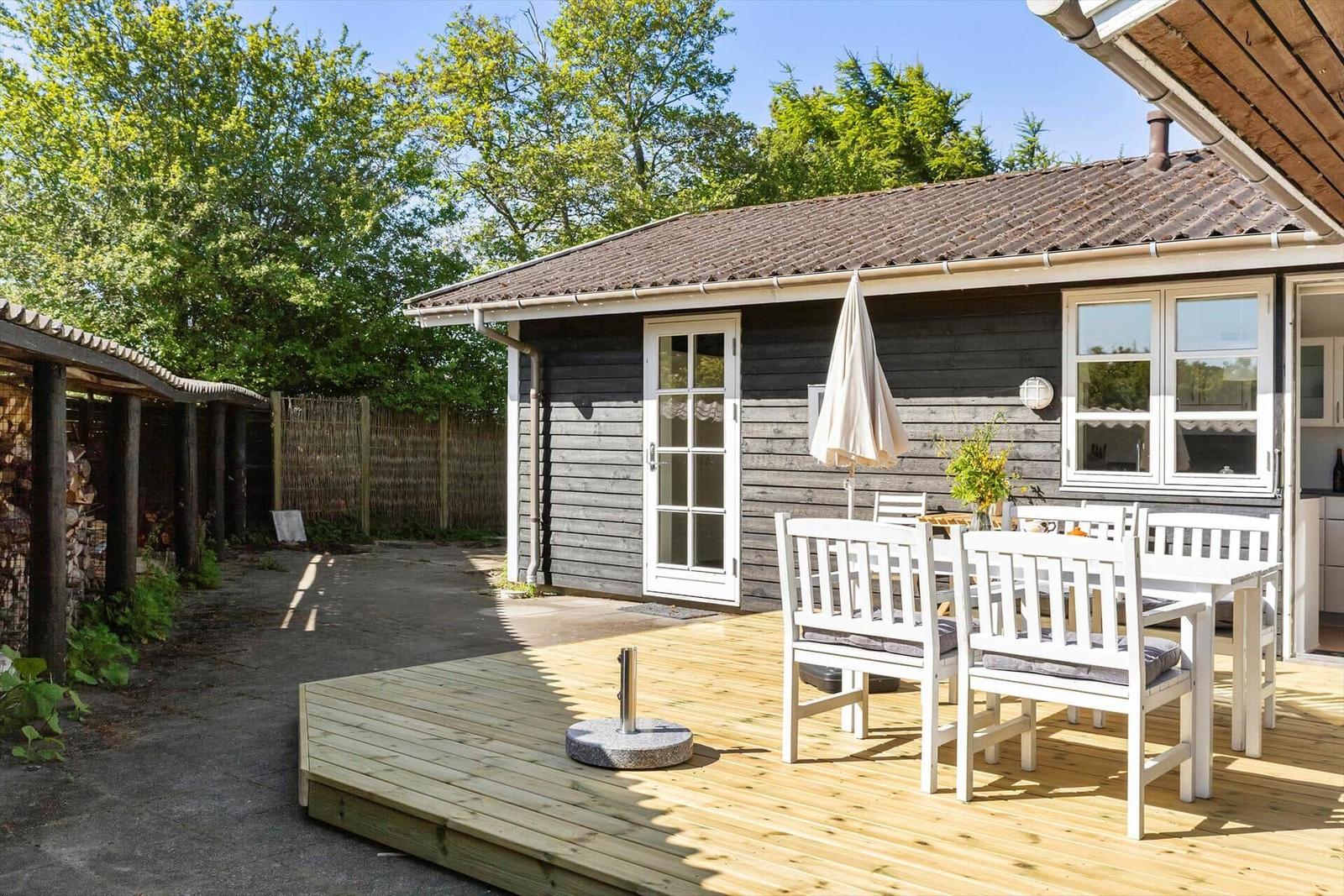 Haus mit Terrasse, weißem Tisch und Stühlen, geschlossenem Sonnenschirm und Holzwand.