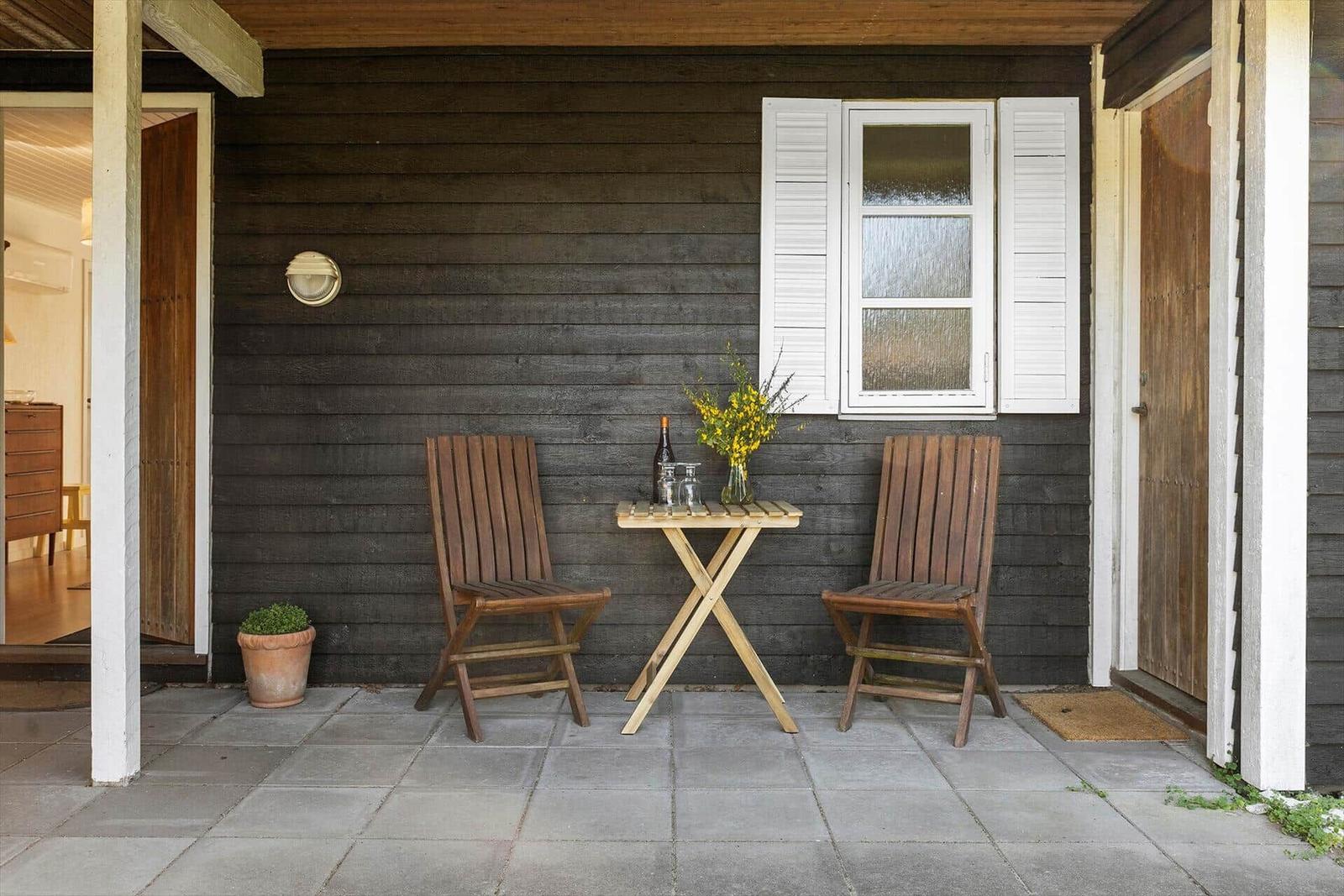 Terrasse mit Tisch, zwei Stühlen, Blumen und Weinflasche vor dunklem Holzhaus.