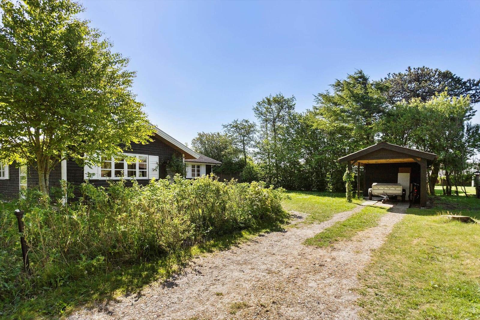 Ein Haus mit dunklem Holz und weißem Fensterrahmen. Ein Holzcarport mit einem Anhänger. Gras und Bäume.