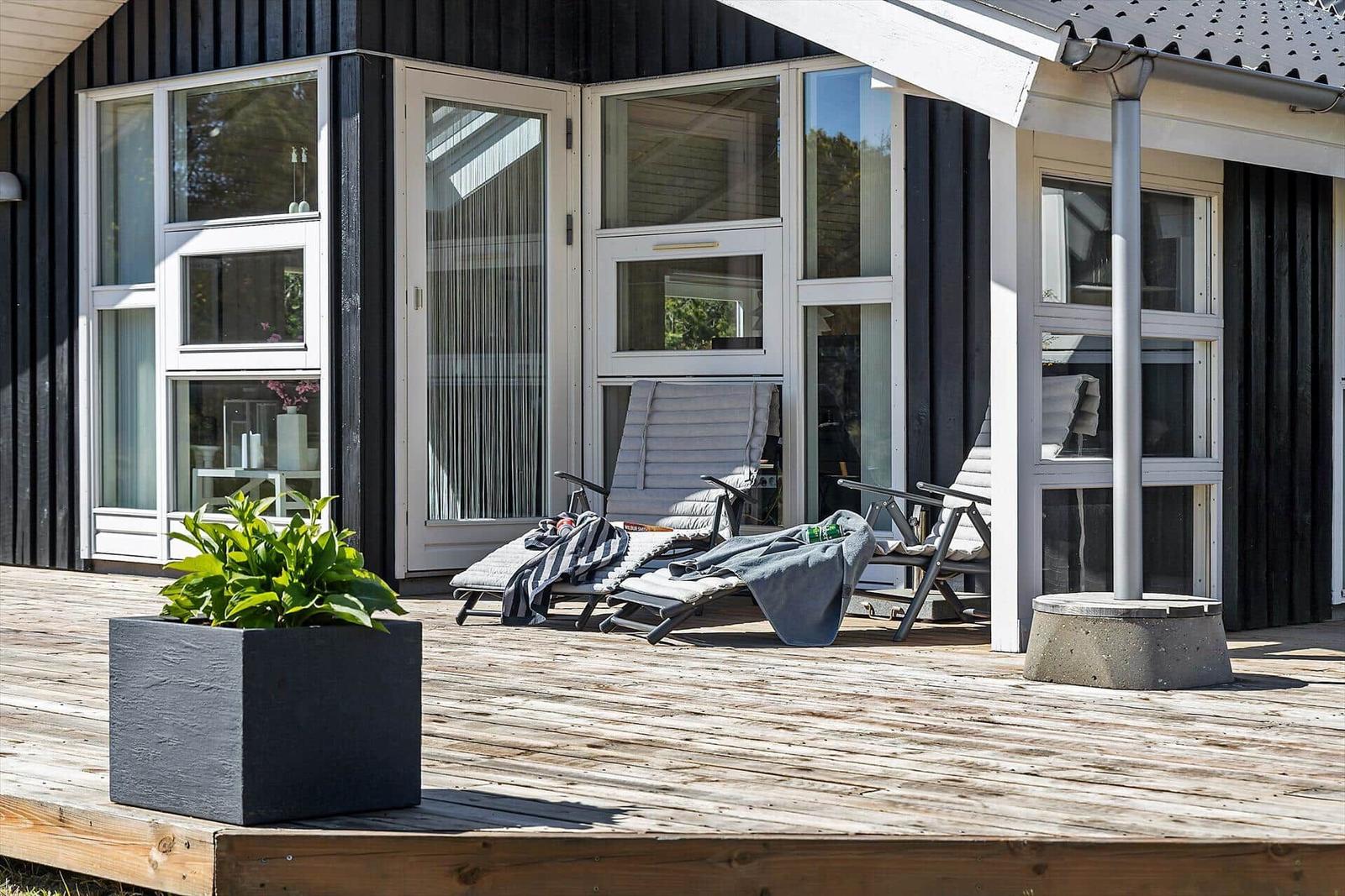 Terrasse mit Liegestühlen und Pflanze vor schwarzem Holzhaus mit großen Fenstern.