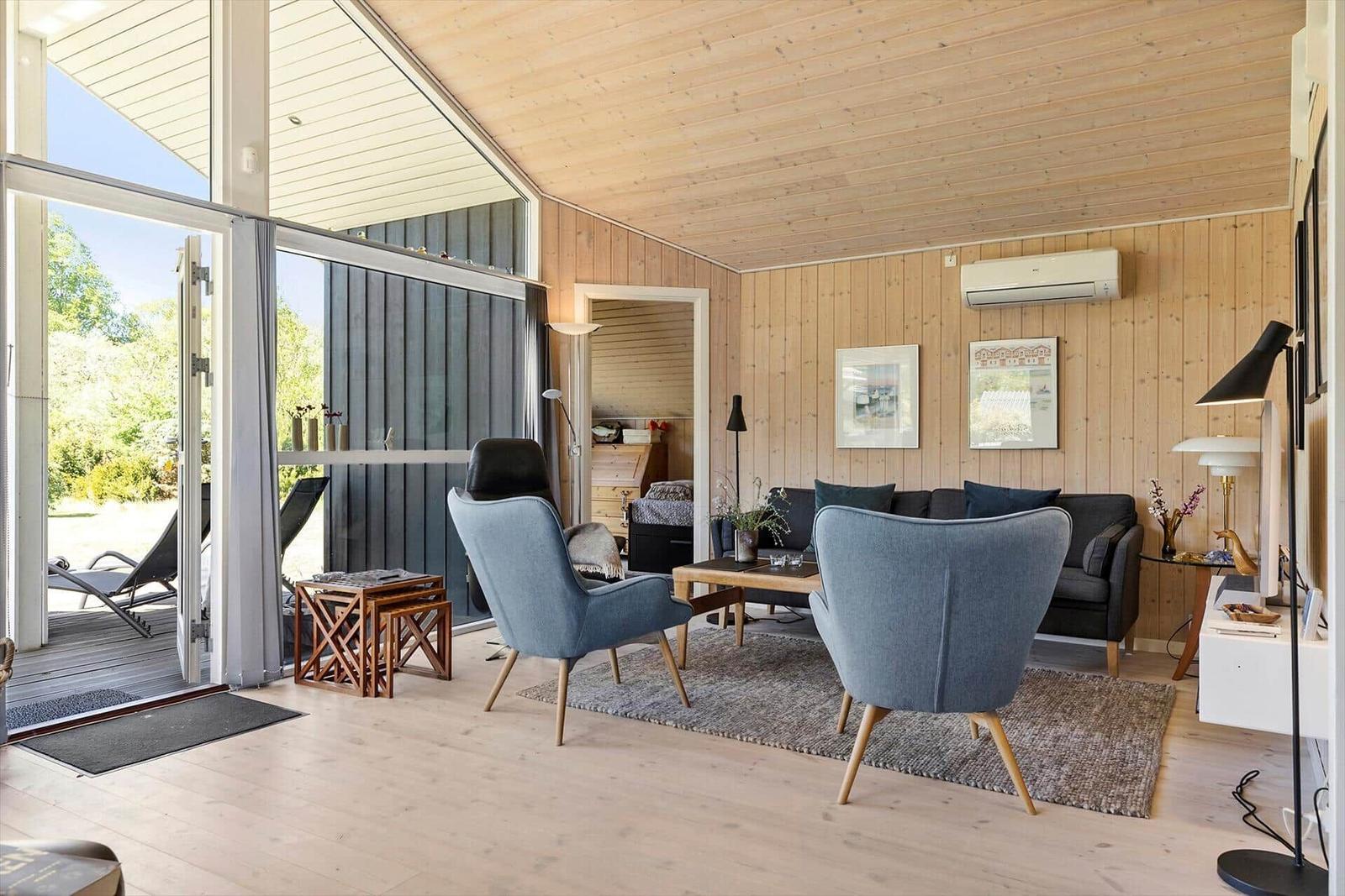 Living room with wooden walls and ceiling, blue chairs and sofa. Entrance to terrace with sun loungers.