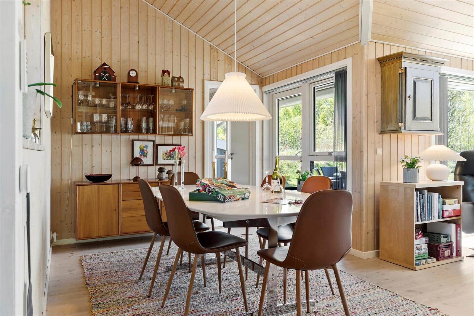 Dining room with wooden walls, dining table and chairs, windows and shelves.