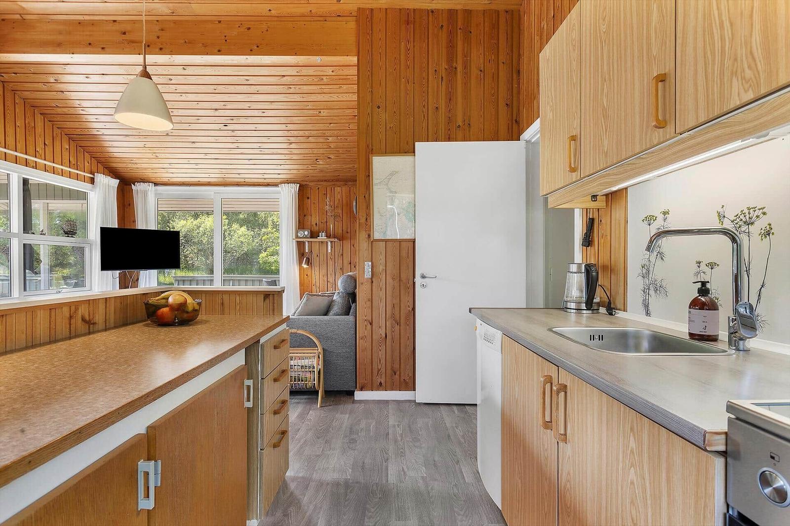 Kitchen with wooden cabinets and view into living area with sofa and windows.