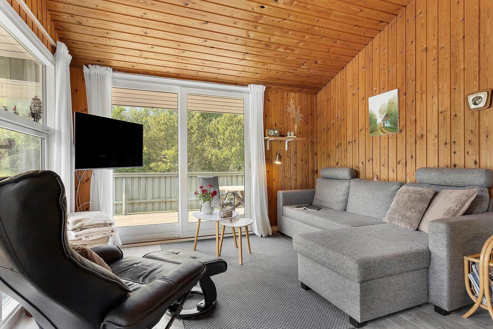 Living room with wood walls, sofa, recliner, and TV. Balcony with view of greenery.