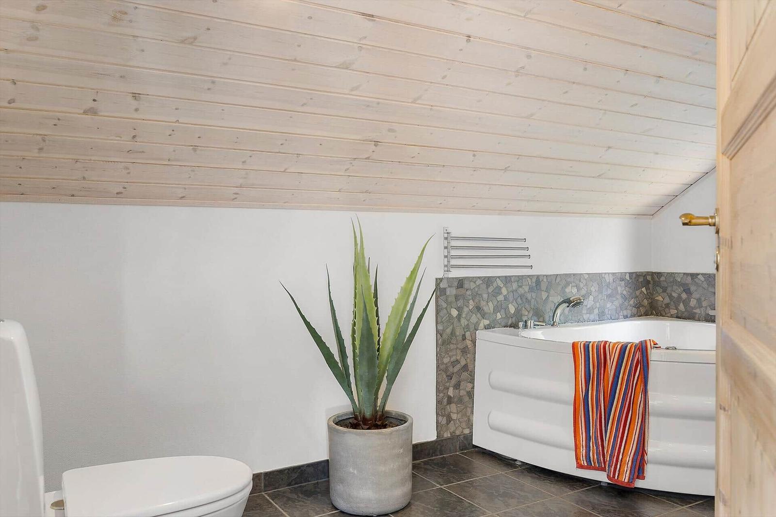 Badezimmer mit Badewanne, Toilette und Pflanze unter Holzdecke.