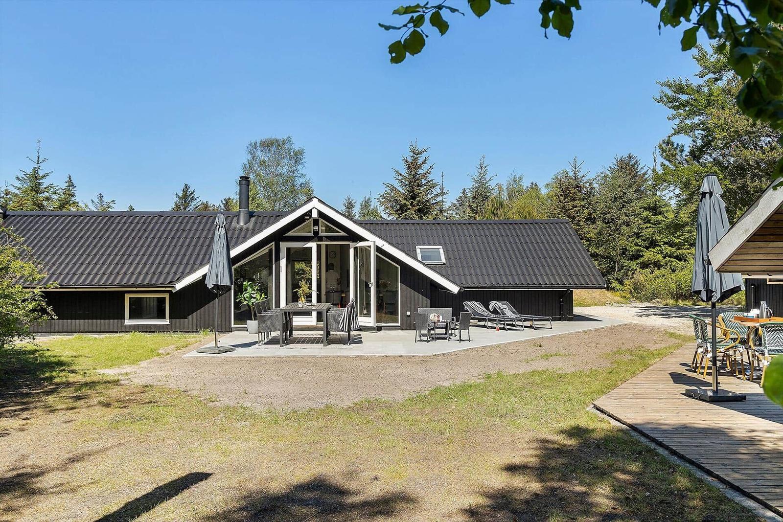 Moderne Holzhütte mit Terrasse, Liegen und Garten. Umgeben von Wald und klarem Himmel.