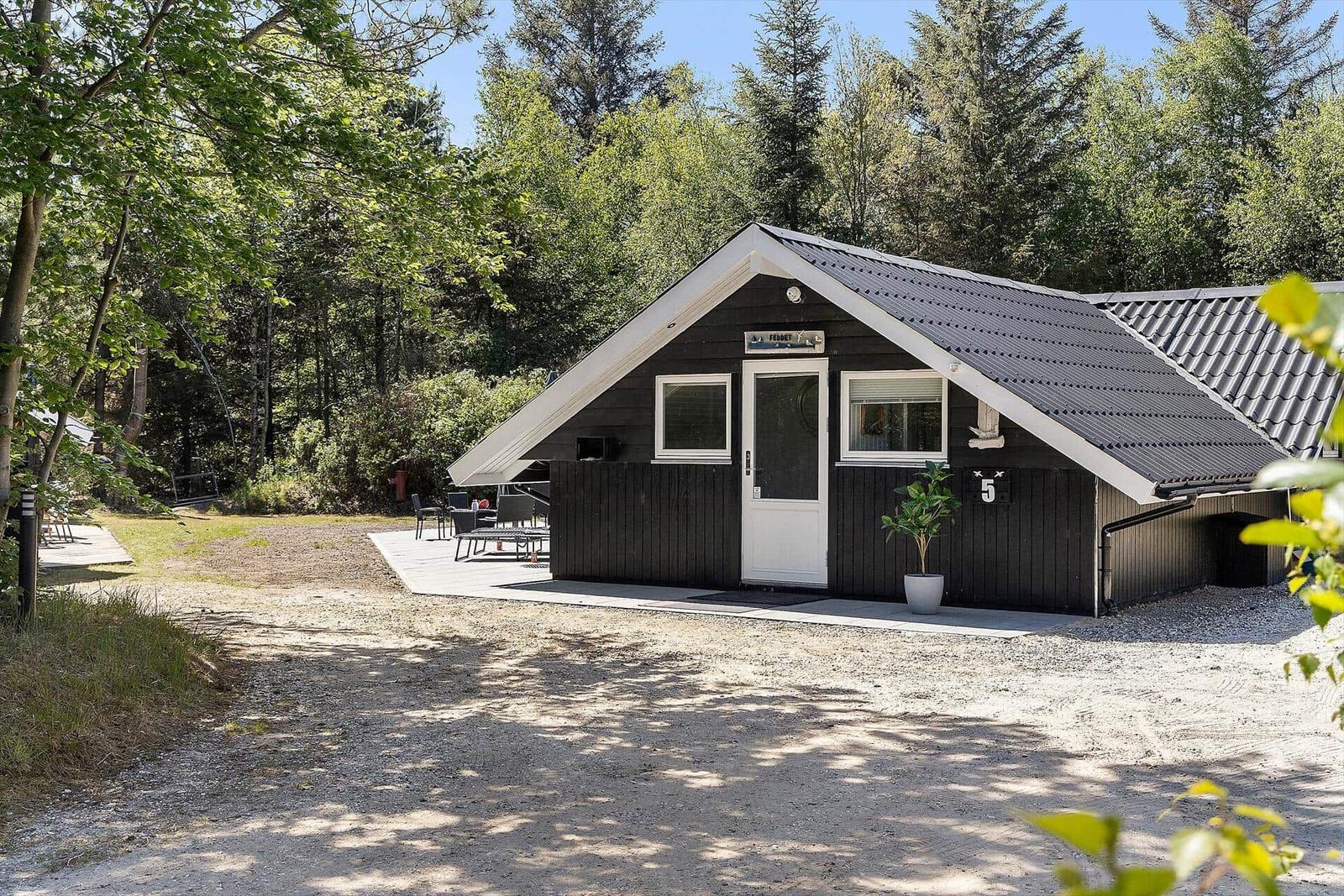 Ein schwarzes Holzhaus mit weißer Tür und Dachrand. Vorhof mit Tisch und Stühlen. Umgeben von Bäumen.