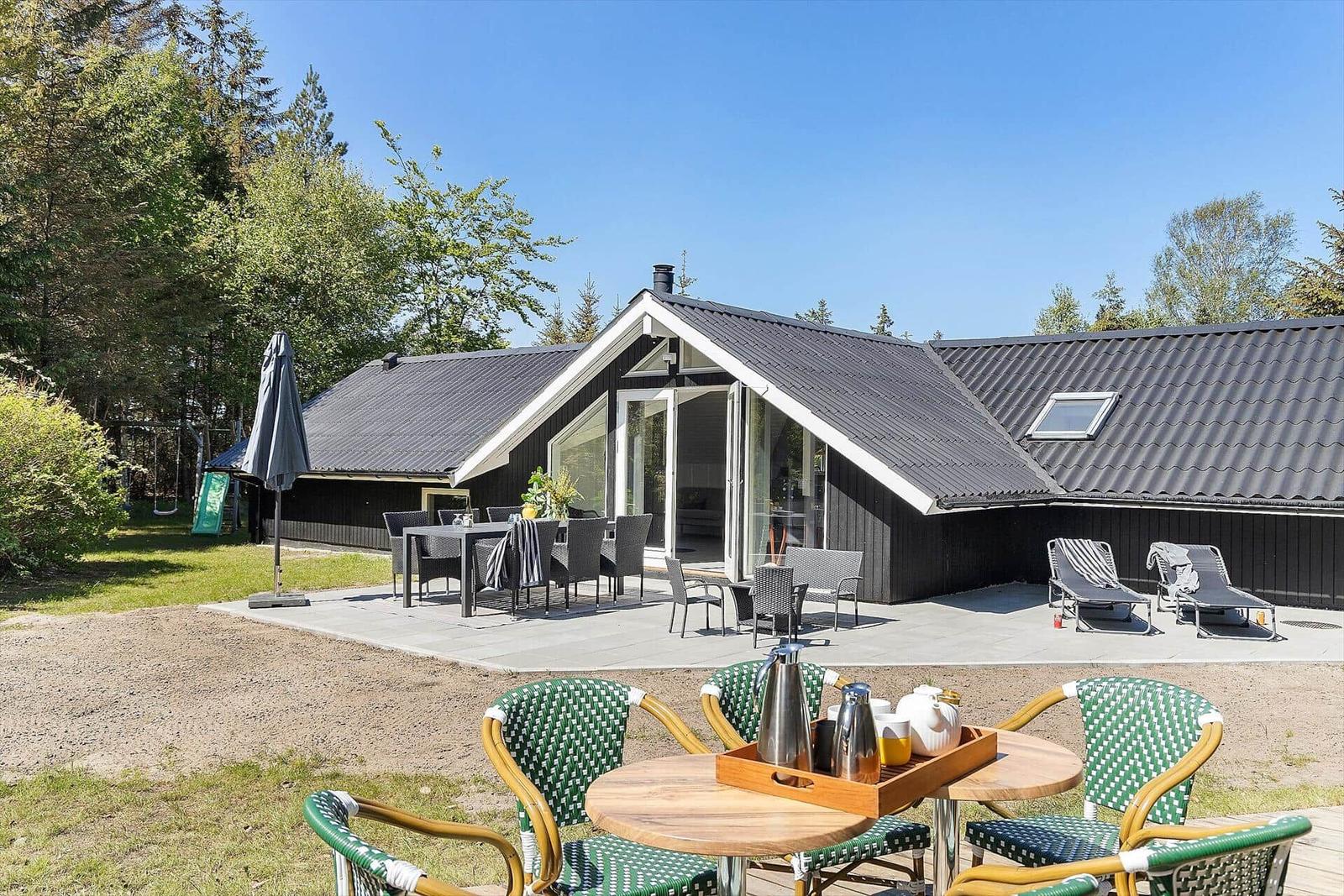 Modernes Haus mit Terrasse, Gartenmöbeln und Liegestühlen. Grüne Bäume umgeben die Anlage.