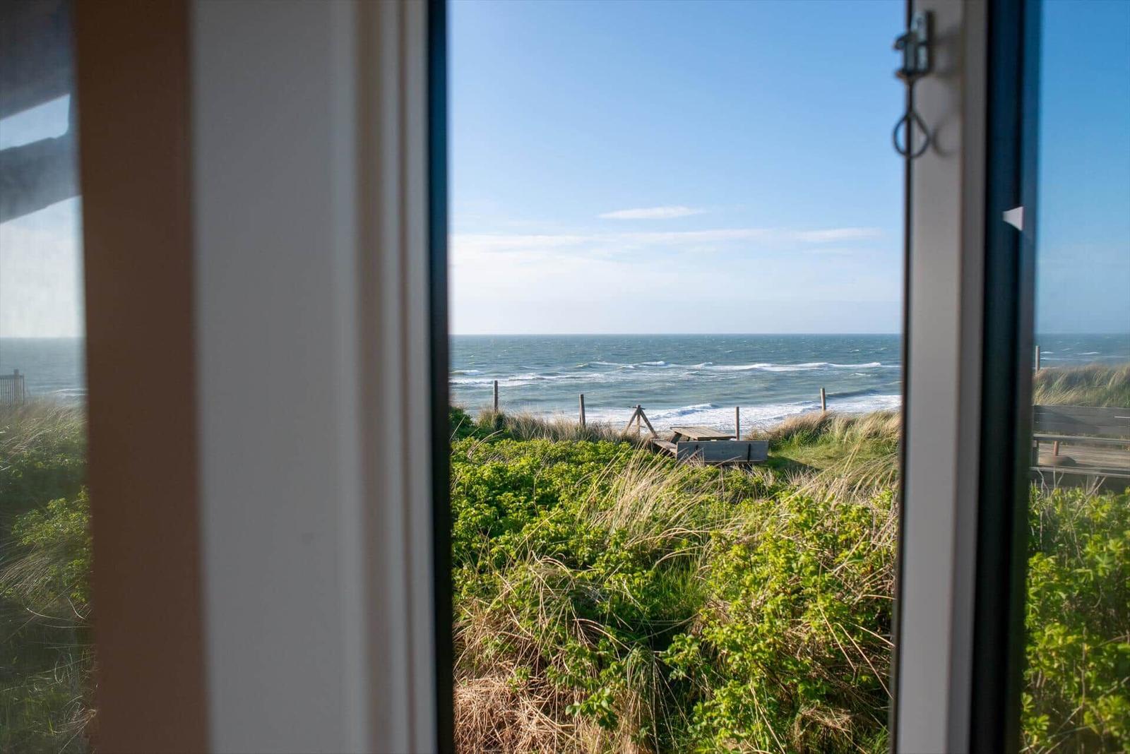 Durch das Fenster blickt man auf das Meer und grüne Dünen.