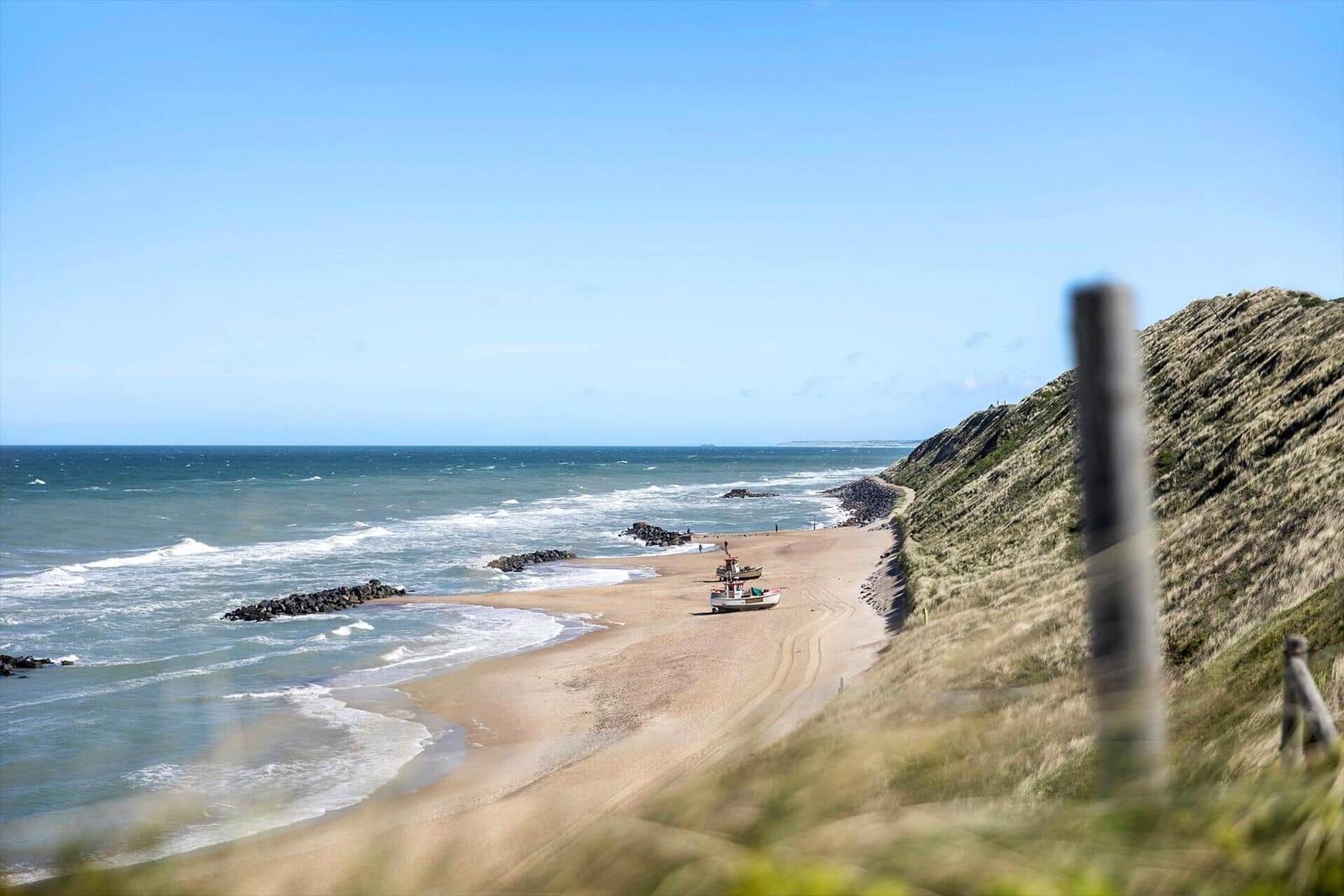 Strand mit Fischerboot, Dünen und Meer unter blauem Himmel.