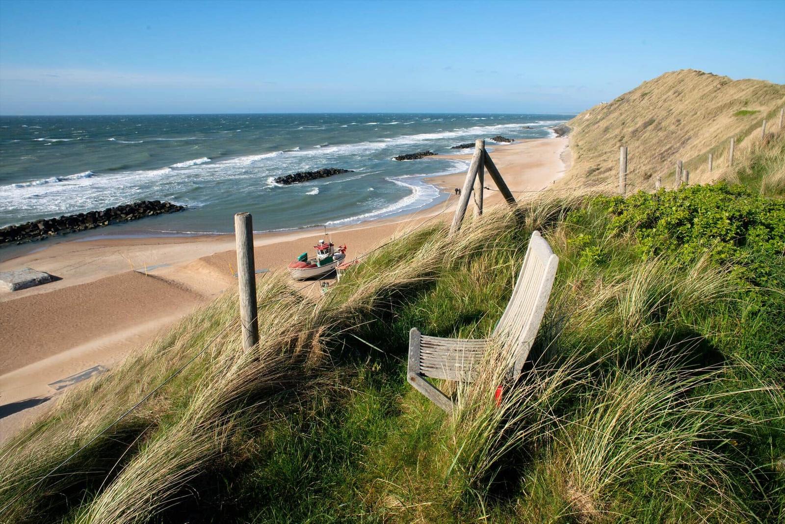 Ein Holzstuhl auf Dünen mit Blick auf Strand und Meer.