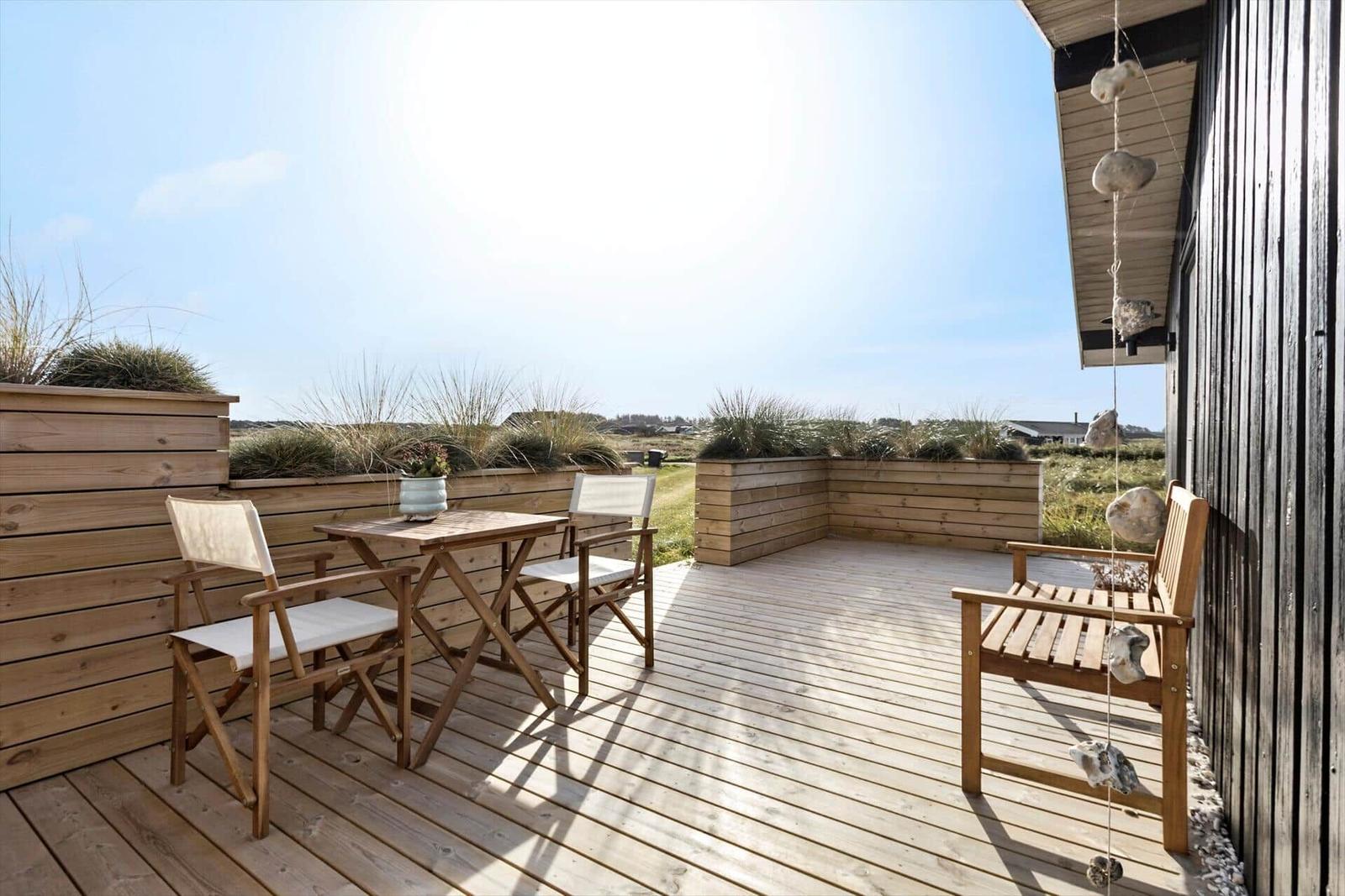 Wooden terrace with table, chairs, and plants. View of grassland and sky.