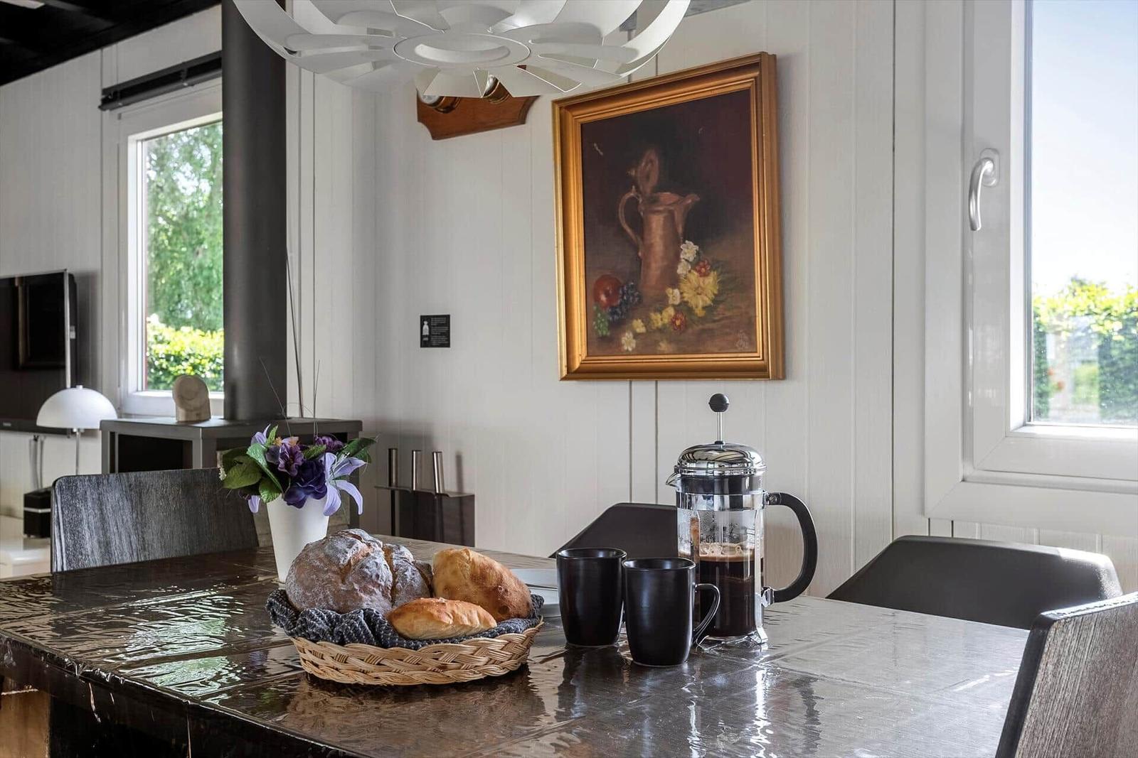 Dining table with basket of bread, coffee maker, and flowers against wall with painting.