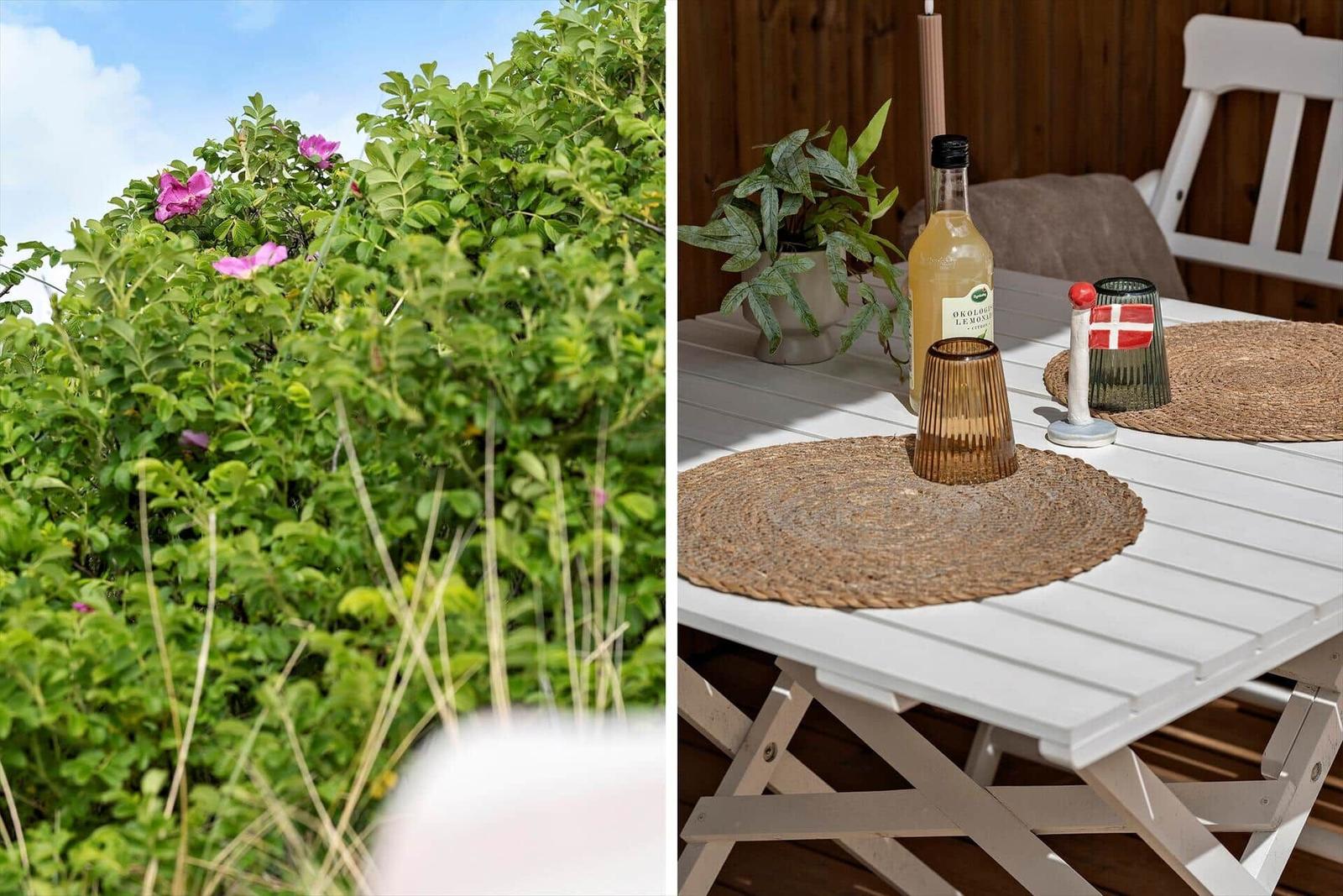 Außenbereich mit Tisch, Stühlen und Pflanzen. Rosa Blüten und Holzwand im Hintergrund.