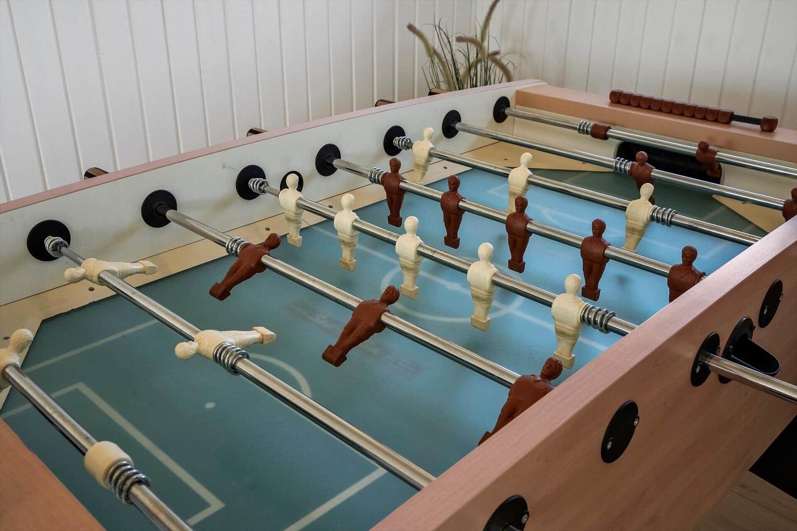 Fussballtisch mit Holzrahmen und Metallstangen. Hintergrund: weiße Wand und Pflanze.
