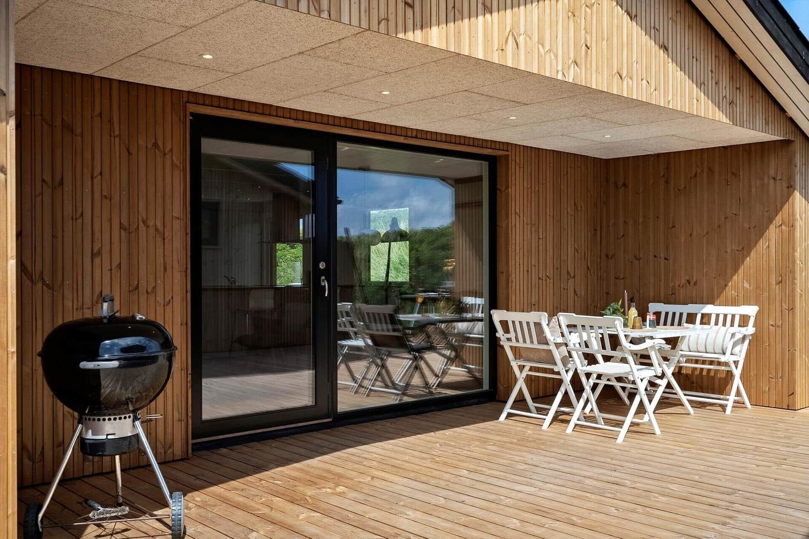 Terrasse mit Grill, weißem Tisch und Stühlen, Holzwand und Glastüren.