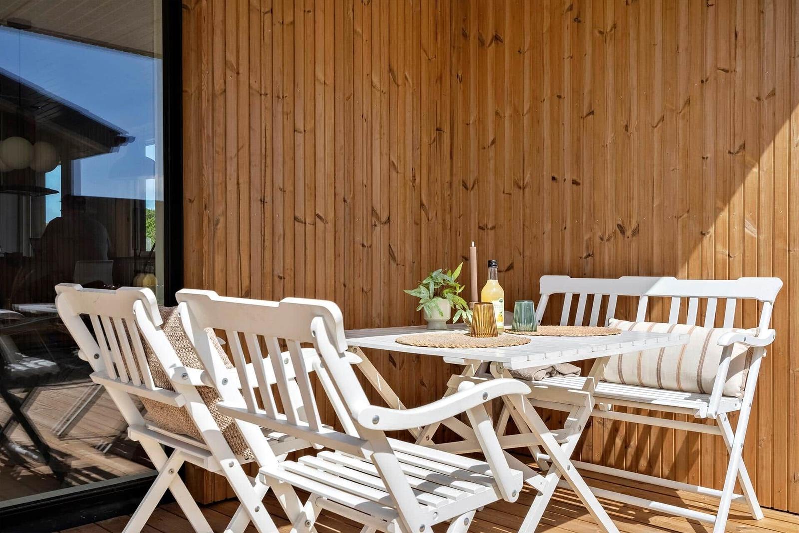 Terrasse mit weißem Tisch und Stühlen, Holzwand im Hintergrund, Sonne scheint.