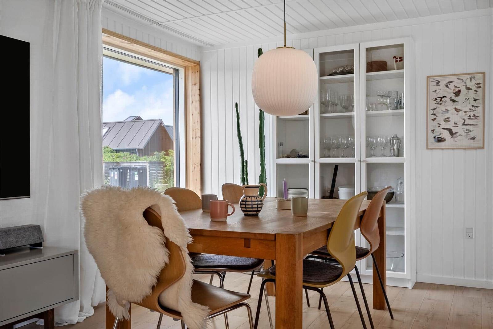 Esszimmer mit Holztisch, Stühlen und Glaswand. Fenster mit Blick auf Dach. Deckenleuchte und Wandbild.