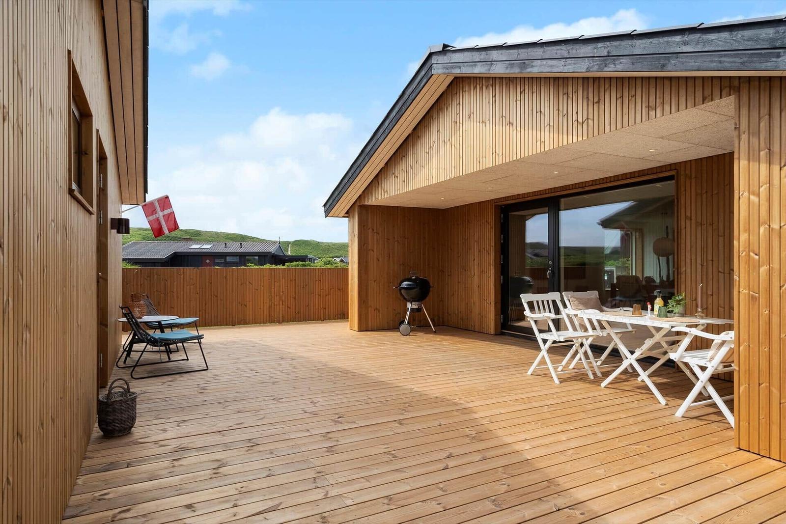 Holzterrasse mit Grill, Tisch und Stühlen. Dänische Flagge weht im Wind.