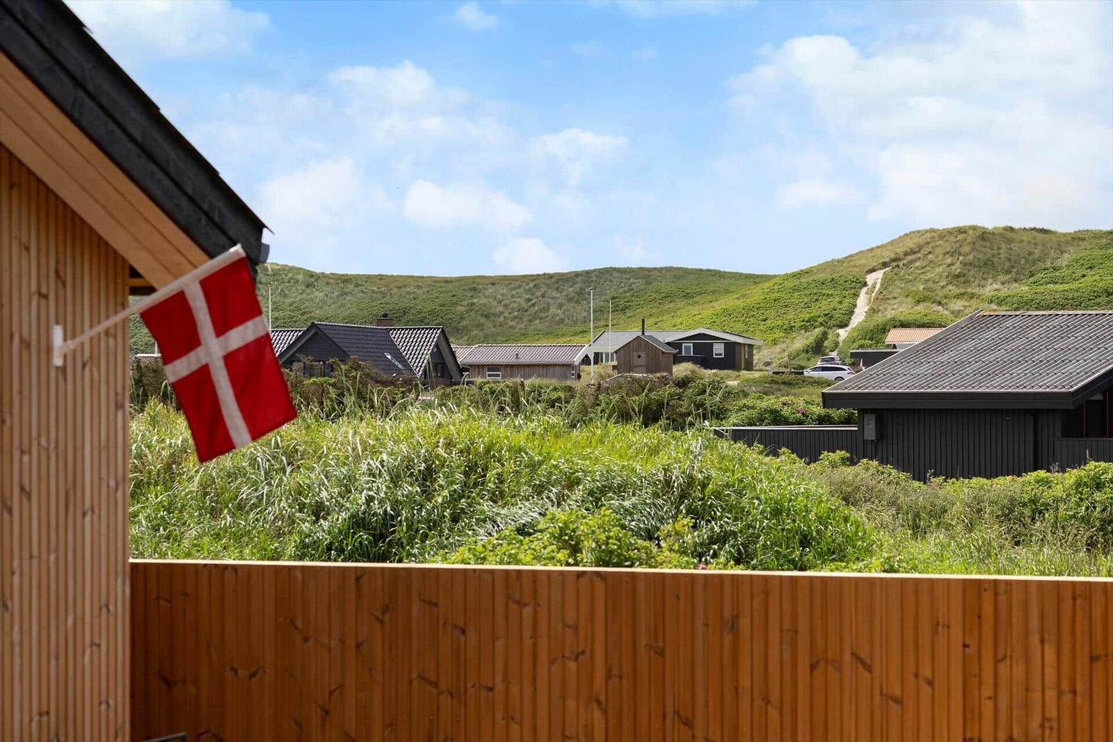 Dänische Flagge weht vor einem Holzzaun mit Blick auf grüne Dünen und Häuser.