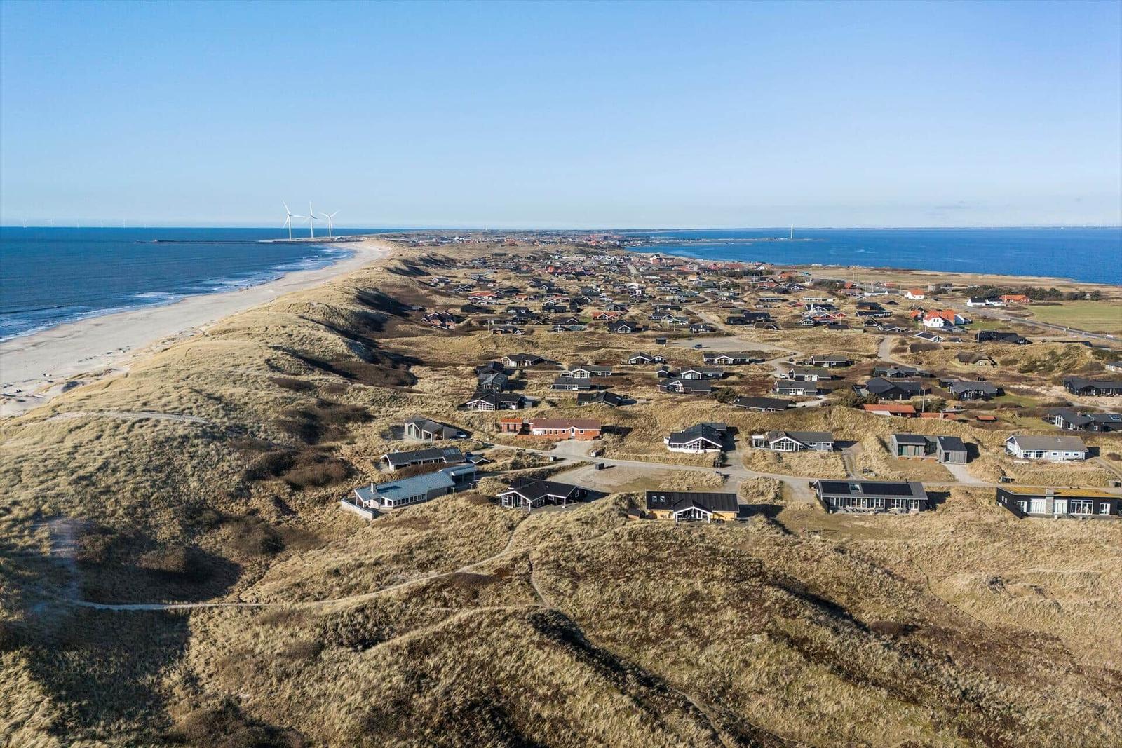 Aerielansicht von Strandhäusern auf Dünen mit Windkraftanlagen am Horizont.