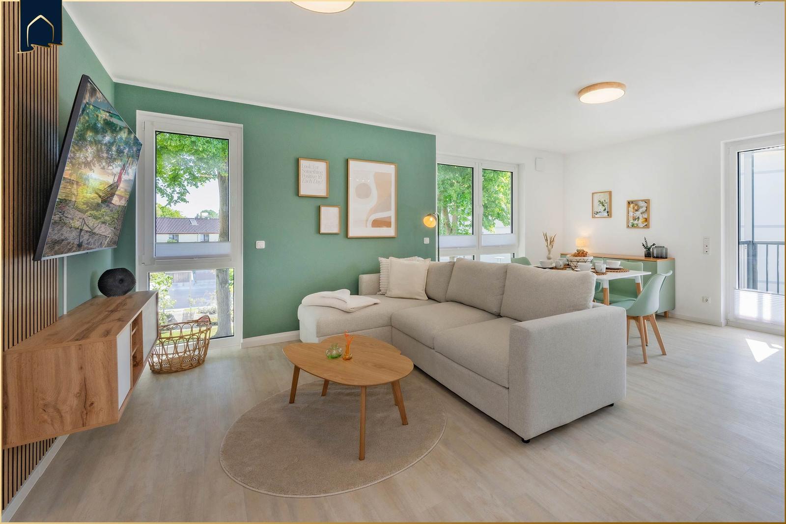 Wohnbereich mit Sofa, Couchtisch und TV. Essbereich mit Tisch und Stühlen. Grüne Wand und Fenster.