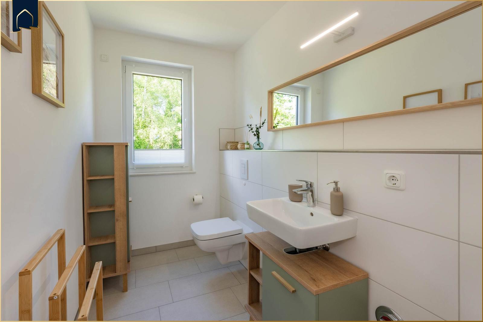 Badezimmer mit Waschbecken, Spiegel, Toilette und Fenster mit Blick auf Grün.