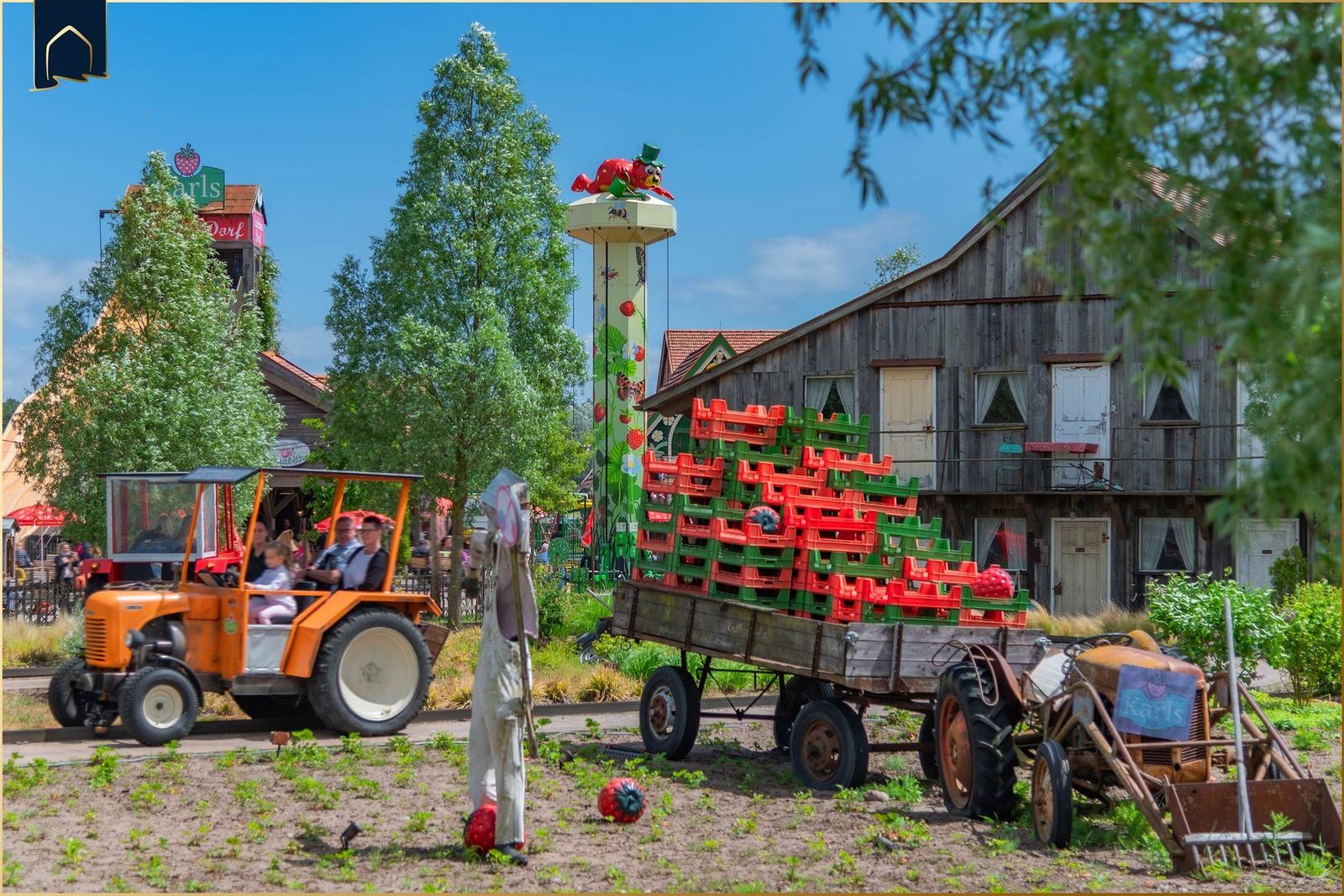 Traktor mit Anhänger voller roter und grüner Kisten vor einem Holzhaus.