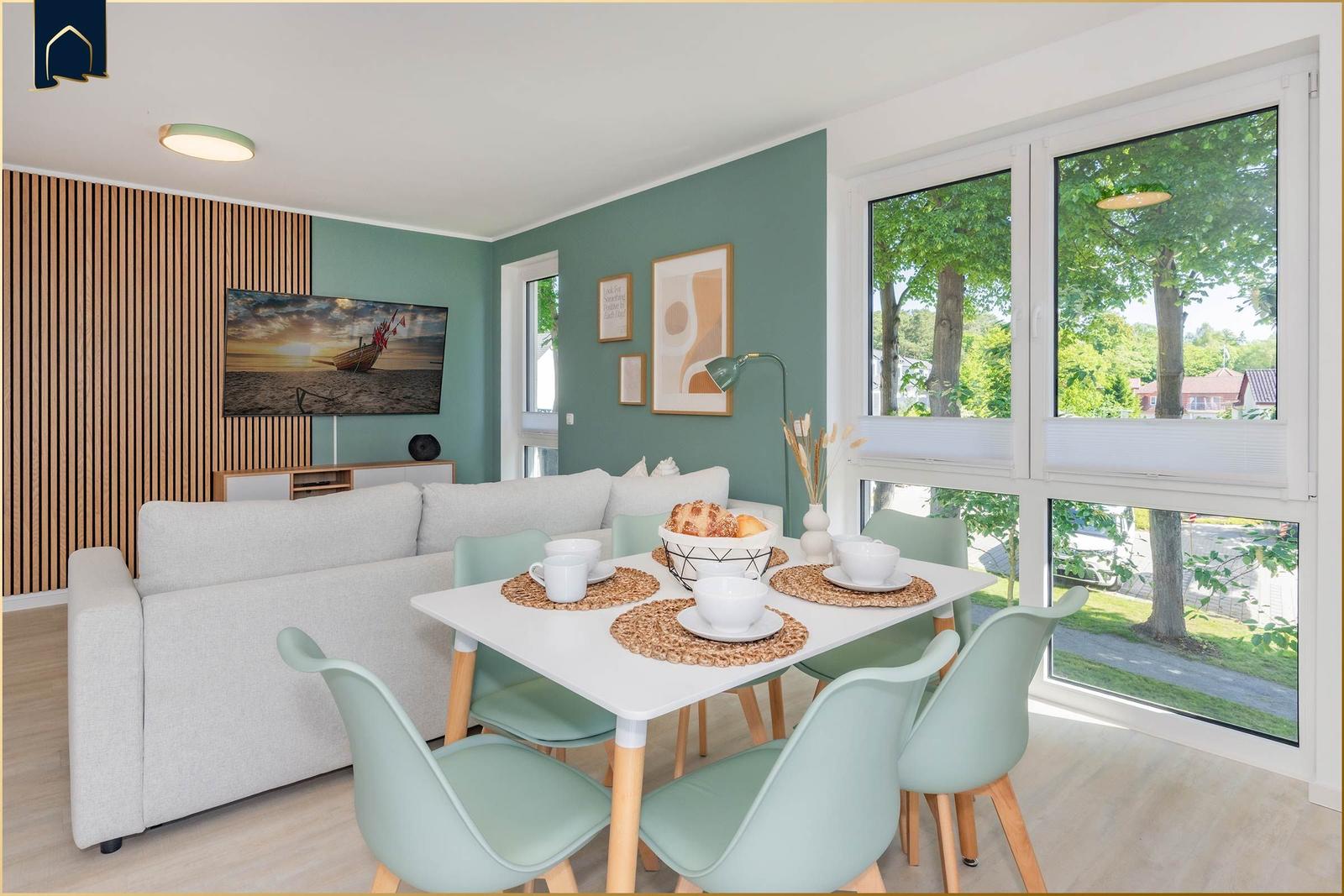 Wohn- und Essbereich mit Sofa, Tisch und Stühlen. Große Fenster mit Blick auf Bäume.