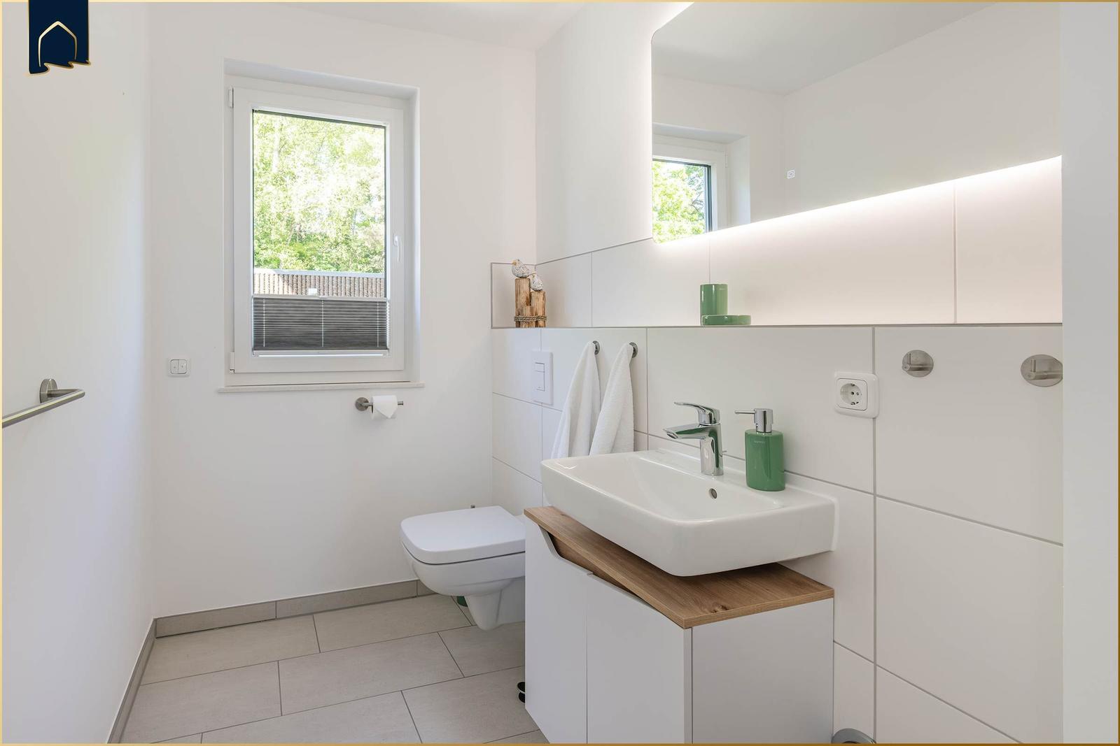 Badezimmer mit Waschbecken, Toilette und Fenster. Spiegelschrank mit beleuchteter Oberseite.