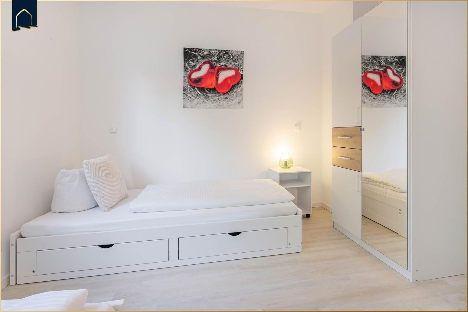 Schlafzimmer mit weißem Bett, Spiegelschrank und Wandbild mit roten Herzen.