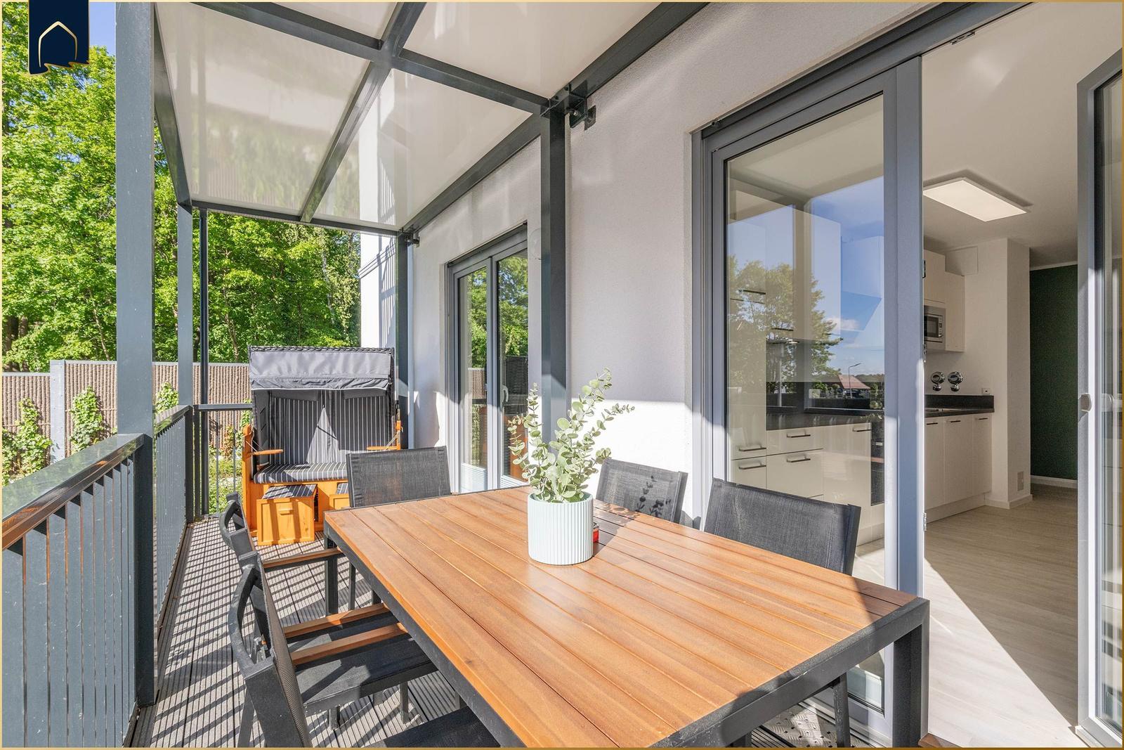Terrasse mit Holztisch, Stühlen und Blick auf grüne Bäume. Durchgehende Glaswände führen ins Haus.