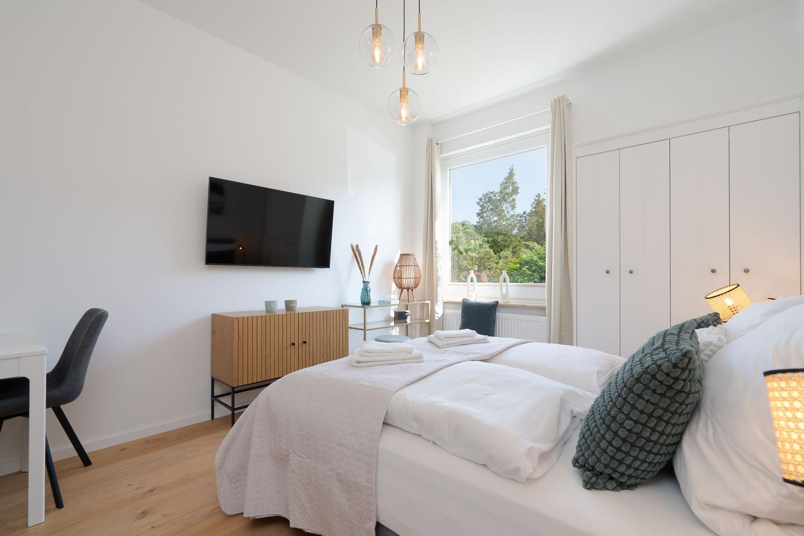 Schlafzimmer mit Bett, TV, Schreibtisch und Fenster mit Blick auf Grün.