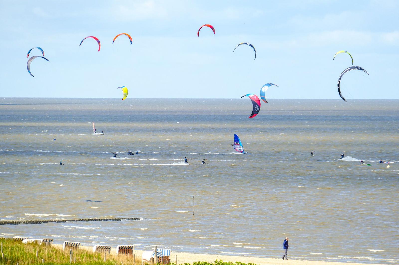 Kitesurfer und Windsurfer genießen den Strand. Mehrere Kites schweben über dem Wasser.