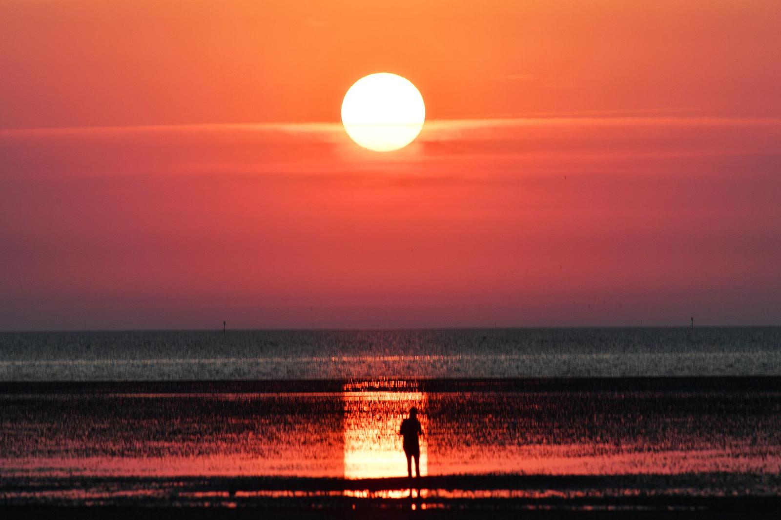 Eine Person steht am Strand und blickt auf einen großen Sonnenuntergang über dem Meer.