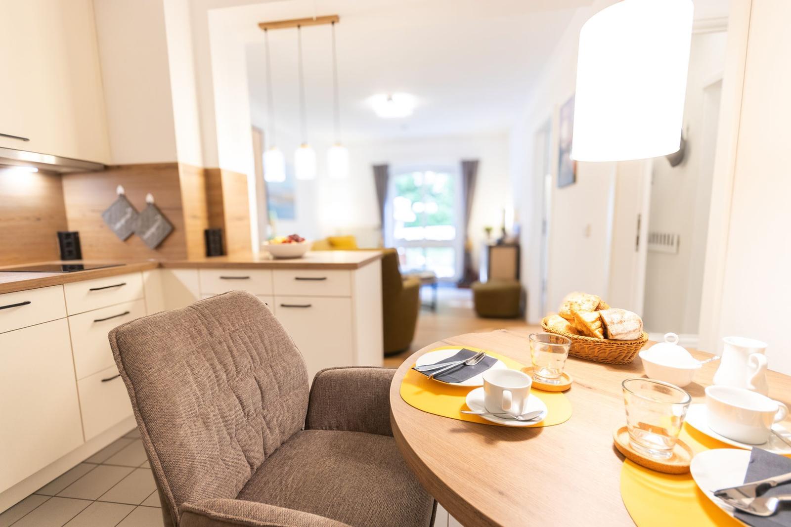 Esstisch mit Stuhl, Kaffeetasse und Brotkorb. Küche und Wohnbereich im Hintergrund.