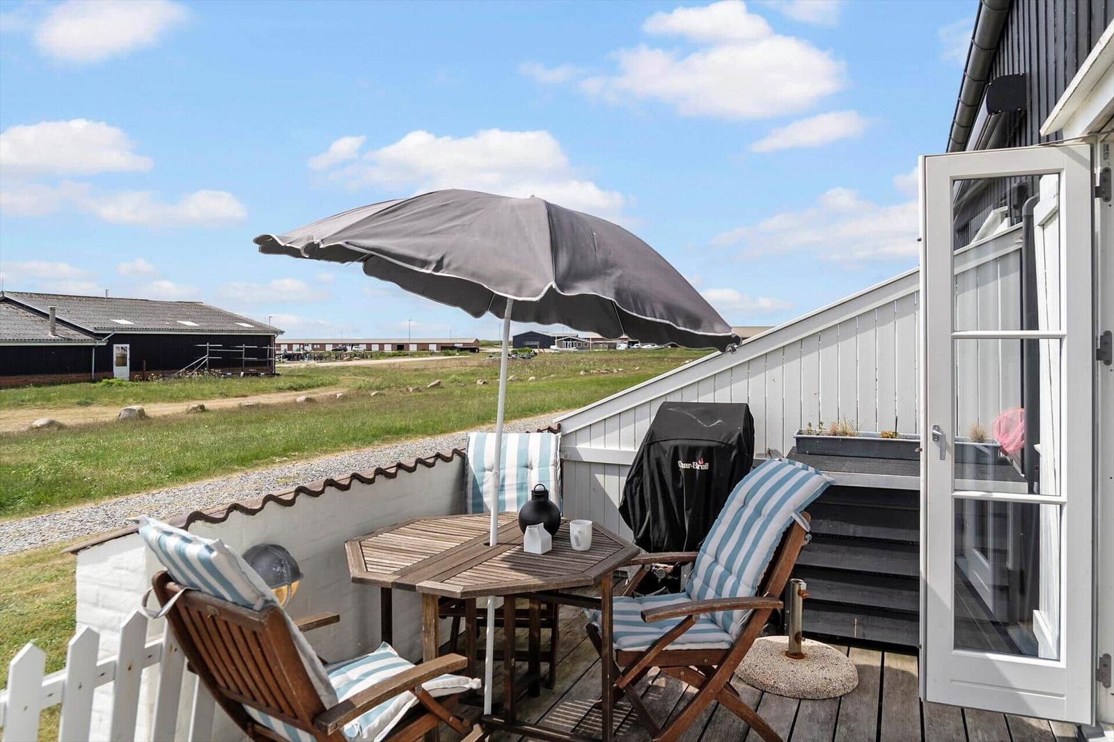 Terrasse mit Tisch, Stühlen und Sonnenschirm. Blick auf grünes Feld und Gebäude.