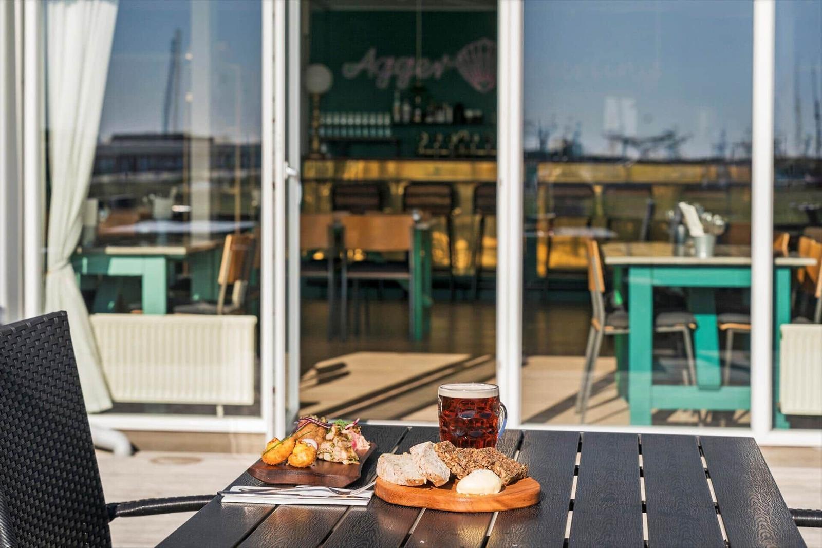 Außenbereich mit Tisch, Getränken und Speisen. Im Hintergrund Restaurant mit Glasfront.